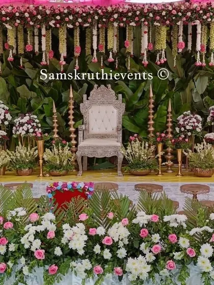 A grand stage setup with a banana leaf backdrop. The decor is enhanced with an ornate silver throne, traditional brass lamps, and a lush border of white and pink flowers.