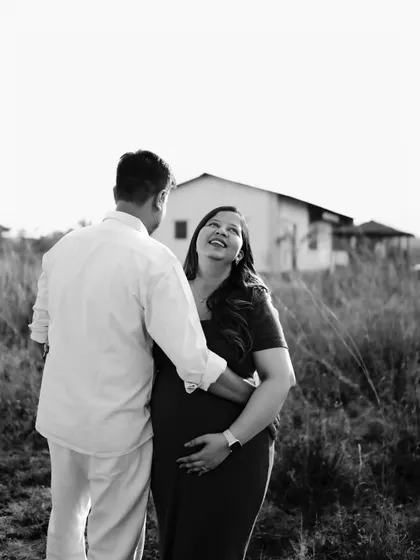 A beautiful black and white portrait of the couple. He embraces her from behind as they share a loving look, with a rustic house in the background.