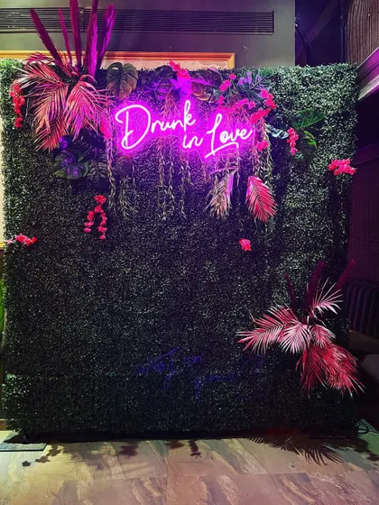 A trendy and edgy photo wall for a cocktail party or sangeet. A 'Drunk in Love' pink neon sign is set against a lush green wall, decorated with tropical pink leaves for a bold and fun statement.