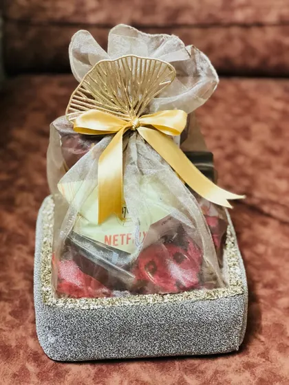 An elegant Rakhi hamper in a grey glittery basket, wrapped in sheer fabric with a gold bow and fan detail.