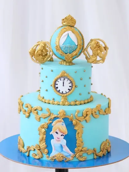 A fairytale Cinderella cake that is pure magic. This two-tiered blue cake is adorned with ornate gold details, a clock striking midnight, and Cinderella's pumpkin carriage as a topper.