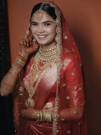 The bride, adorned in a stunning red saree and gold jewelry, smiles for the camera, radiating happiness and traditional elegance.