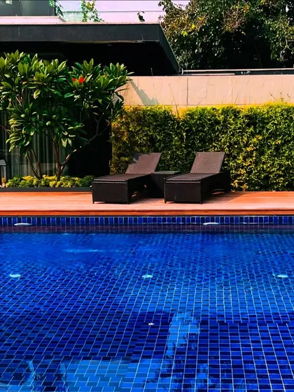 The swimming pool area at the Sultanpur farmhouse, complete with a wooden deck and comfortable lounge chairs. The space is framed by a tall green hedge for privacy, creating a serene and exclusive oasis.