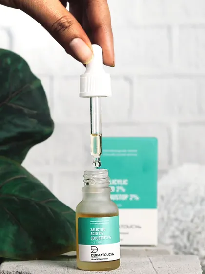 This action shot shows the dropper of a Dermatouch face serum, highlighting the product's liquid consistency and ease of application against a natural, leafy backdrop.