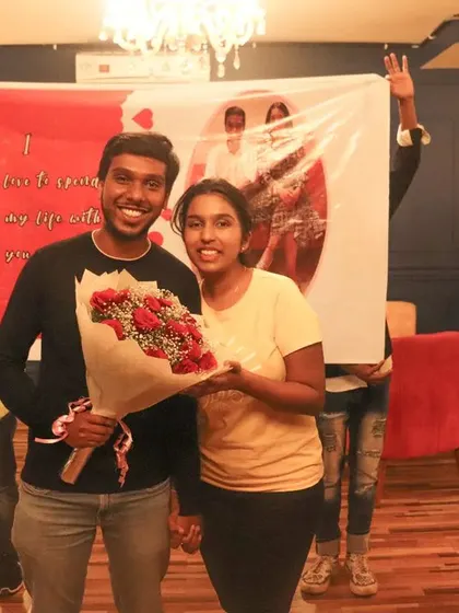 A happy couple poses with a bouquet of roses after a successful surprise proposal. Our team holds up a banner that reads "I would love to spend the rest of my life with you" to complete the moment.