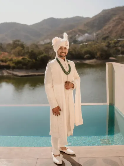 The groom, looking dapper and serene against a beautiful poolside backdrop. His classic white sherwani and green necklace exude understated elegance.