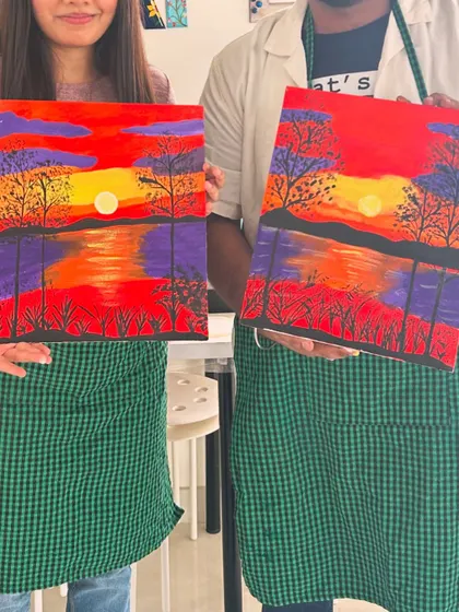 A couple holding their matching sunset landscape paintings.