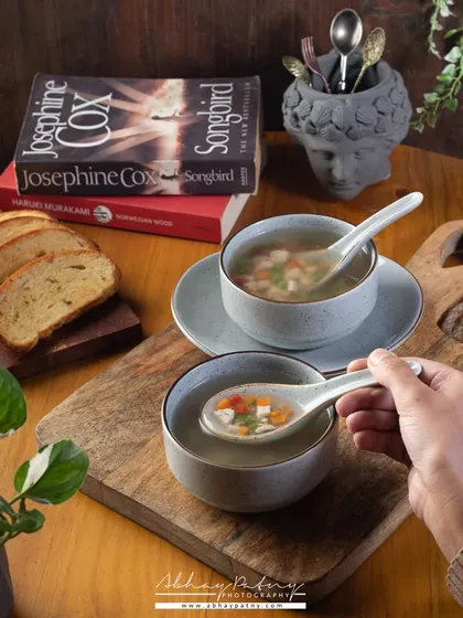 This soup shot is styled to feel warm and comforting, perfect for a cozy cafe. The inclusion of a person's hand and books creates a relatable, lifestyle-oriented scene.