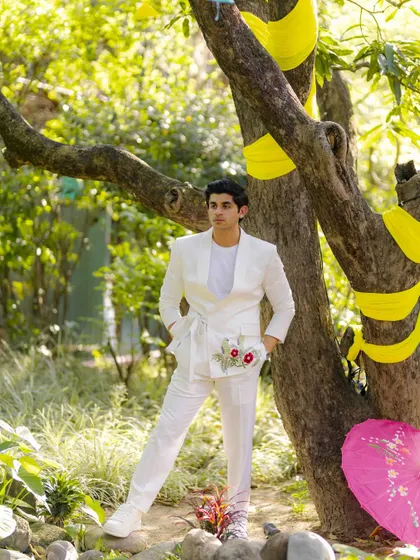 A stylish portrait of the groom at an outdoor Haldi or Mehendi event. His modern white outfit with floral embroidery stands out against the lush green background.
