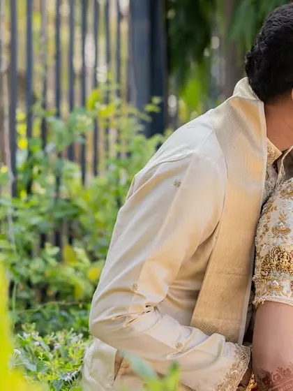 A tender, candid moment of affection captured during their heritage photoshoot. The soft natural light and lush green foliage add a touch of softness to the traditional setting.
