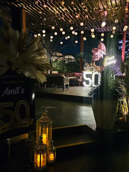 The entrance to a stylish 50th birthday party. The welcome sign and lanterns lead guests to a beautiful rooftop setting with string lights and a glamorous photo backdrop in the distance.