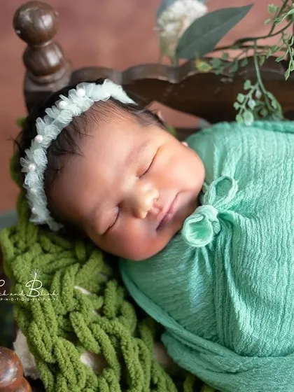 A sweet and adorable close-up. The focus is on the baby's sleeping face, framed by a mint green swaddle and a delicate headband.