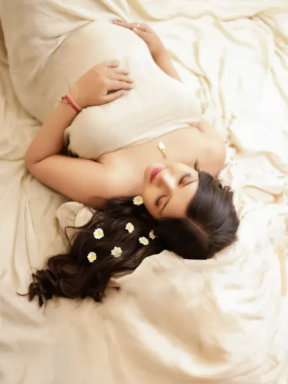 A peaceful and serene overhead shot. The mom-to-be lies on a soft, neutral-colored fabric, with delicate daisies in her hair.