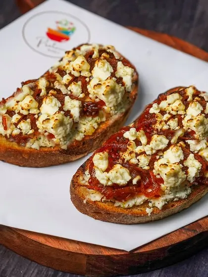 Two pieces of bruschetta with a savory topping of feta cheese and onion jam, served on a wooden board. The dark background creates a cozy, rustic ambiance perfect for an Italian eatery.