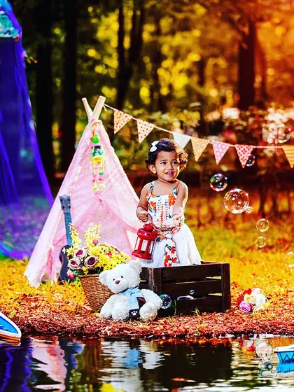 A toddler girl enjoys a magical picnic by the water, complete with a teepee, bubbles, and toy boats. This elaborate outdoor setup creates a fantasy world for her to play in.