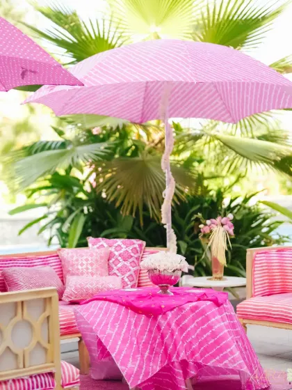 A chic lounge area from our 'Serene Vibes' Mehendi. A pink leheriya-print umbrella provides shade for a seating arrangement with matching tie-dye fabrics and delicate floral centerpieces.