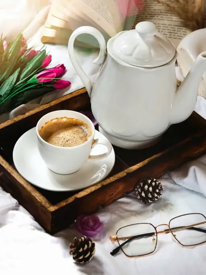 A cozy morning scene with a cup of coffee on a wooden tray in bed. The props, like the open book, glasses, and flowers, help tell a story of a slow, relaxing start to the day.