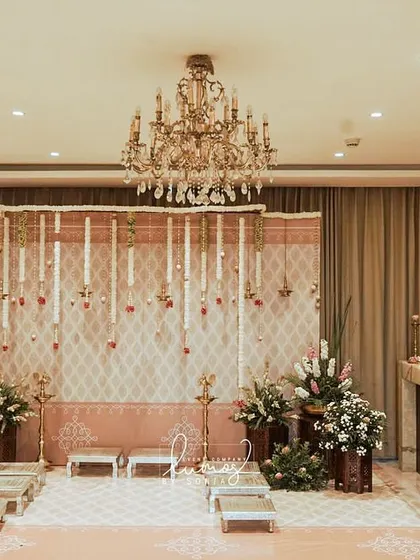 An intimate and traditional engagement setup inside a home. The decor features a backdrop of hanging jasmine garlands and elegant brass lamps, creating a warm and sacred ambiance.