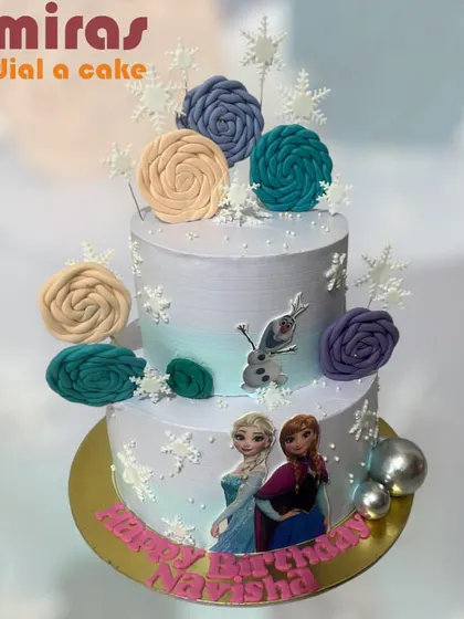 A magical two-tier cake for a Frozen fan, featuring edible images of Anna and Elsa, a fondant Olaf, and beautiful frosted rosettes. The delicate snowflakes and shimmering silver balls complete this winter wonderland creation.