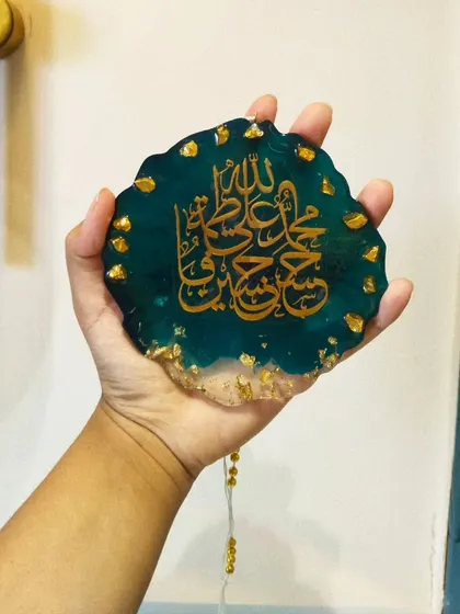 This is a custom resin car hanging featuring beautiful Islamic calligraphy. This piece includes the Panjatan names in gold against a deep teal and clear resin base, accented with gold flakes for an elegant finish. It's a meaningful accessory for your car's interior.