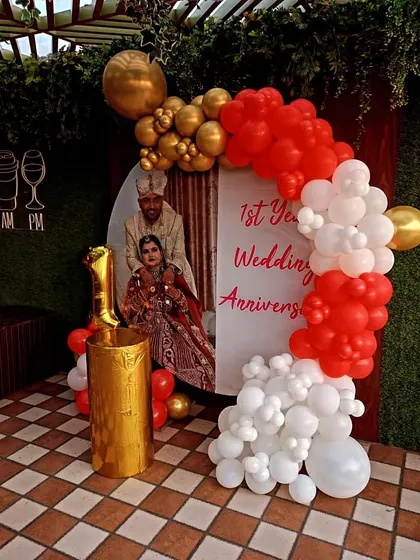A personalized first wedding anniversary setup with a custom photo backdrop and a red, white, and gold balloon arch.