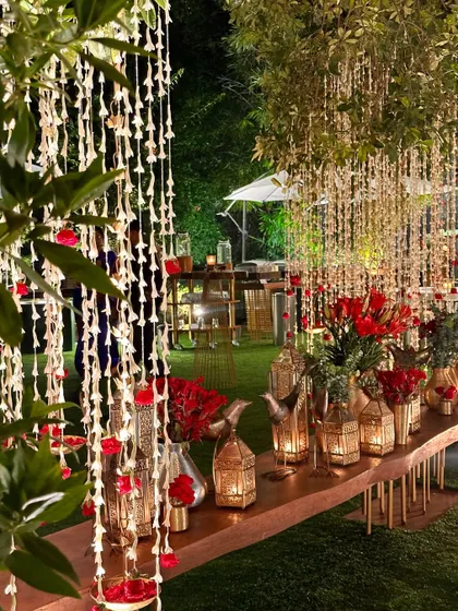 A close-up of a table decorated with red flowers, golden lanterns, and hanging tuberose strings.