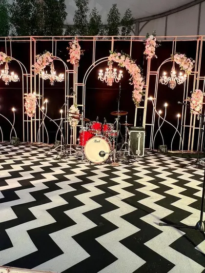 A full view of the elegant stage we set up for the live band. The design features a black backdrop with white metal arches, hanging crystal chandeliers, and soft pink floral arrangements, all set on a bold patterned floor.
