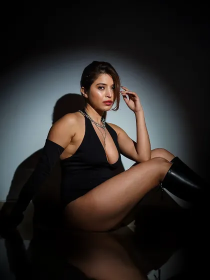 A dramatic, low-key portrait in a black bodysuit and boots. The single spotlight creates a theatrical effect, highlighting the model's form against the darkness.
