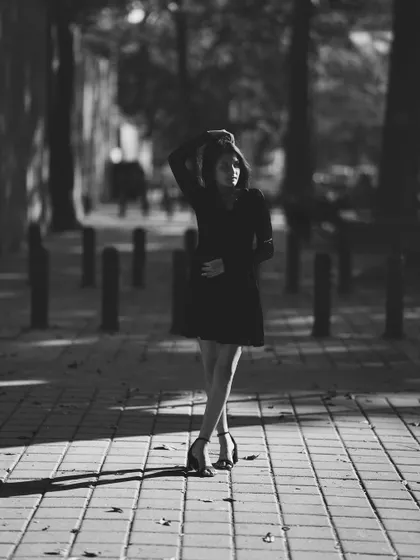 A full-body black and white street portrait of a woman standing on a paved walkway, with dramatic shadows cast by the evening sun. This shot has a strong cinematic and urban feel.