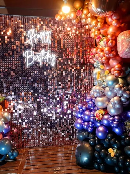 A full view of a dramatic and colorful balloon installation for a birthday party. The organic garland flows from warm oranges and golds to cool silvers, purples, and blacks, creating a striking contrast against the sparkling backdrop.