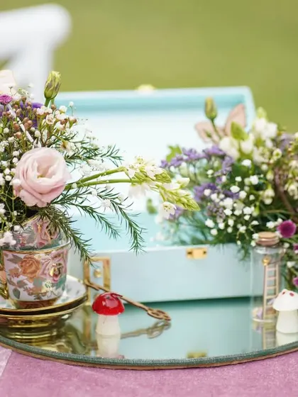 A beautiful centerpiece featuring a teacup overflowing with flowers, set on a mirrored tray with other Wonderland-inspired elements.