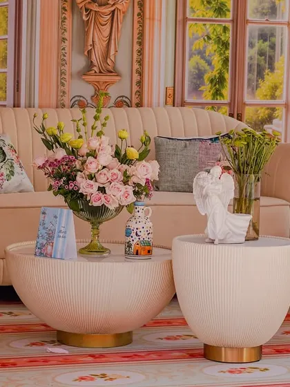 A cozy seating nook from the 'Mint to Be' event, featuring a cream-colored sofa and modern tables. The backdrop is a painted mural of a romantic garden, and the decor includes whimsical angel statues and fresh pink roses.