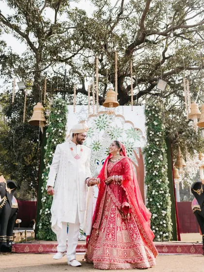 A beautiful shot of the couple walking hand-in-hand away from the mandap, framed by traditional decor and a large tree.