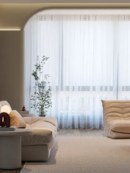 This living room is a study in soft minimalism. The plush, low-profile sofa and armchair are arranged to create an open, inviting feel. Sheer curtains diffuse natural light, and subtle curves in the architecture soften the modern aesthetic.