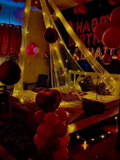 A dimly lit, romantic bedroom decoration. The canopy with fairy lights, red balloons, and heart-shaped balloon pillar creates a very intimate and special atmosphere.