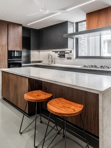 The island in this Ahmedabad kitchen provides a casual seating area, with warm wooden stools complementing the walnut cabinetry and contrasting with the dark island base.