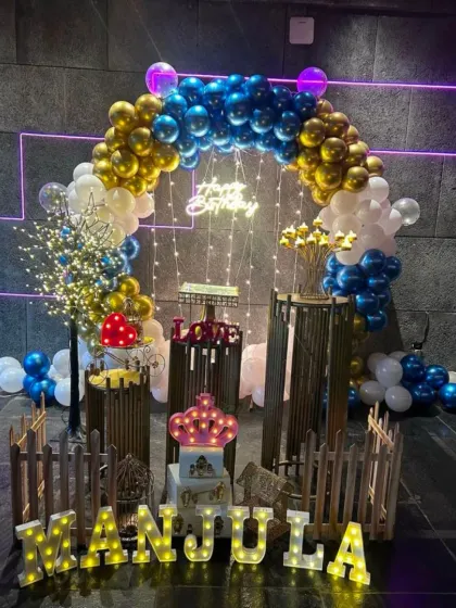 An elaborate party setup with a blue and gold balloon arch, a lighted tree prop, and various decorative items in front of a picket fence.