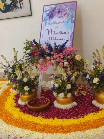 A beautiful welcome setup for a Mehendi ceremony. The personalized sign is surrounded by lush floral arrangements in gold pots and a rangoli made of fresh flower petals, creating an elegant first impression.