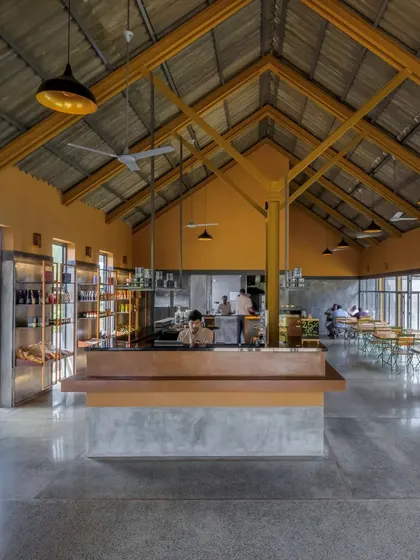 The main hall of Teyla restaurant features a high, vaulted ceiling supported by a metal structure, creating a large, airy expanse. The open kitchen and simple furnishings contribute to a welcoming, chaithela-like atmosphere.