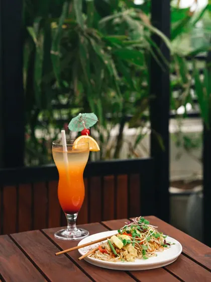 This image pairs a main dish with a refreshing cocktail, creating a complete meal setting. The natural backdrop and wooden table give it a relaxed, al fresco dining vibe.