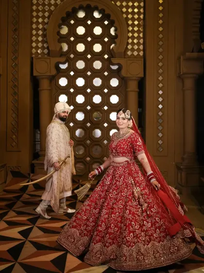A regal portrait of the bride and groom. The bride's magnificent red lehenga is on full display as the groom stands by her side in the palace like corridor.