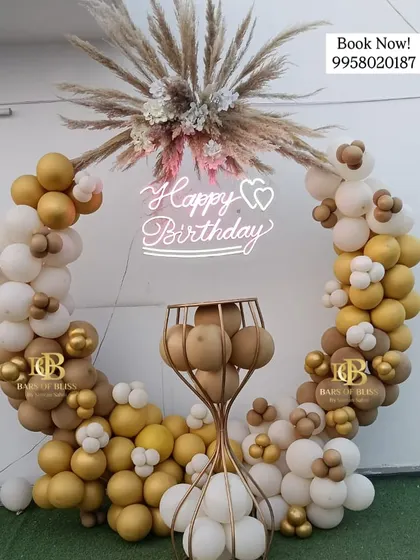 This rustic-chic birthday decor features a full ring of white and gold balloons, topped with a spray of pampas grass. A neon sign and a golden pedestal for the cake complete this elegant setup.