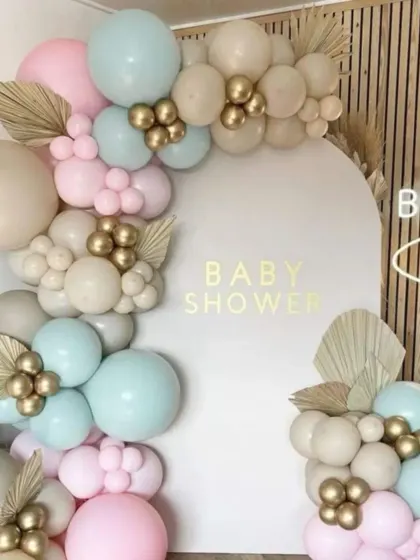 A boho-chic baby shower setup. The pastel blue, pink, and beige balloons are accented with gold clusters and dried palm leaves for a trendy and stylish look.