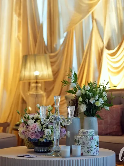 A luxurious lounge area set against a backdrop of hand-painted wallpaper and dramatic golden drapes. The pastel-colored sofas and floral arrangements create a soft, inviting space.