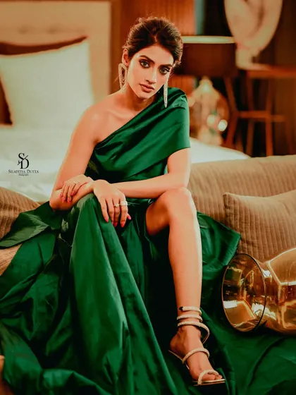 Actress Nusrat Jahan looking elegant in a one-shoulder green satin gown. This full-length shot on a sofa showcases the dramatic silhouette of the dress and her poised, high-fashion pose.