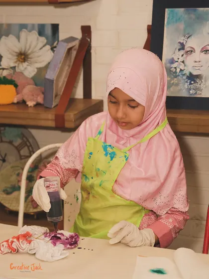 A young girl in a hijab and apron carefully works on her tie-dye project, adding purple dye to her design.