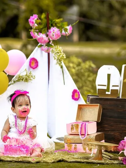 A wide shot of an outdoor cake smash, showing the full picnic-style setup with a teepee, balloons, and decorative suitcases.