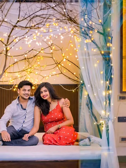 A cozy and imaginative indoor setup featuring a bed with sheer curtains and trees wrapped in fairy lights. This creates a unique, dreamlike setting for a couple's portrait.
