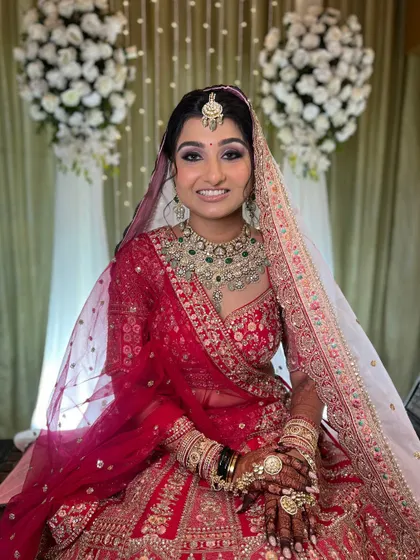 A beautiful bride seated against a floral backdrop. The makeup features a soft purple and pink smokey eye to complement her red lehenga.