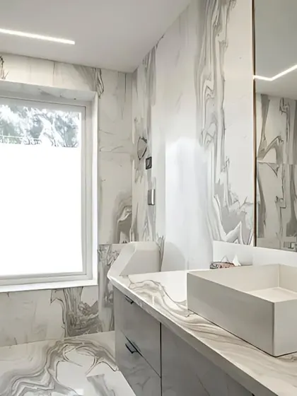 This executive bathroom exemplifies understated luxury with its monochrome palette. The walls and countertop feature Satvario Gold stone with flowing grey veins, creating a cohesive and sophisticated space that is filled with natural light from a large window.
